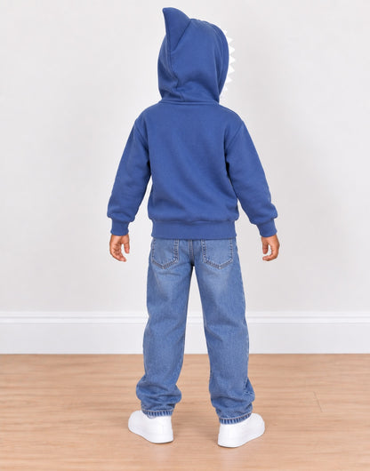 A child standing with their back to the camera, wearing a royal blue hooded sweatshirt with a shark-fin detail on the hood, blue jeans, and white sneakers, set against a plain white wall and light wood floor.