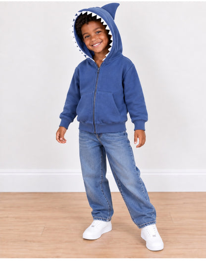 A young child wearing a blue zip-up hoodie with a shark teeth and fin hood, paired with blue jeans and white sneakers, standing on a light wood floor against a white wall.