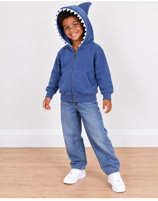 A young child wearing a blue zip-up hoodie with a shark teeth and fin hood, paired with blue jeans and white sneakers, standing on a light wood floor against a white wall.