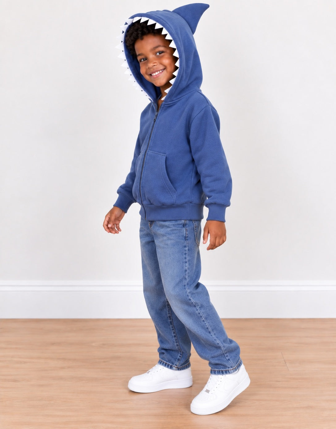 Side view of a young child wearing a blue zip-up hoodie with a shark hood, paired with blue jeans and white sneakers, standing on a light wood floor against a white wall.