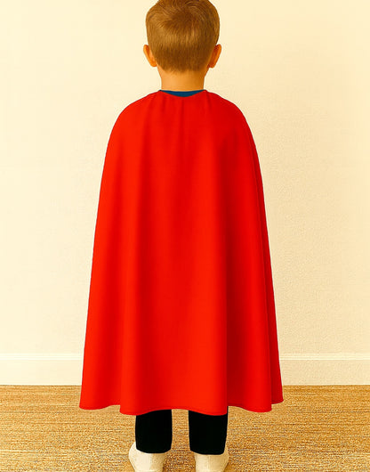 Back view of kid wearing a red cape over a blue shirt, and black pants on a beige background.