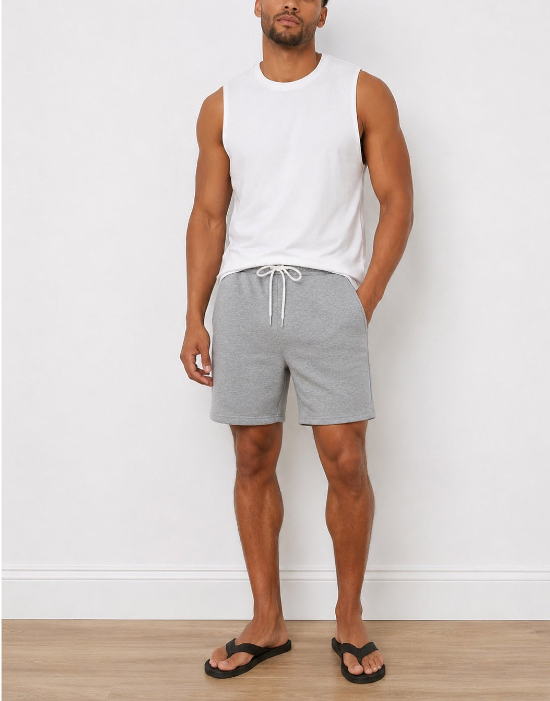 A man modeling light gray heather Drawstring Sweat Shorts with a white sleeveless tank top and black flip-flops, standing against a white wall and light wood floor.