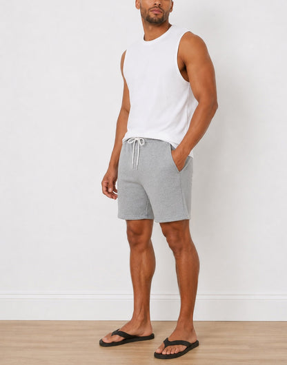 Side view of a man wearing light gray heather Sweat Shorts with a white drawstring, a white sleeveless tank top, and black flip-flops, standing against a white wall.