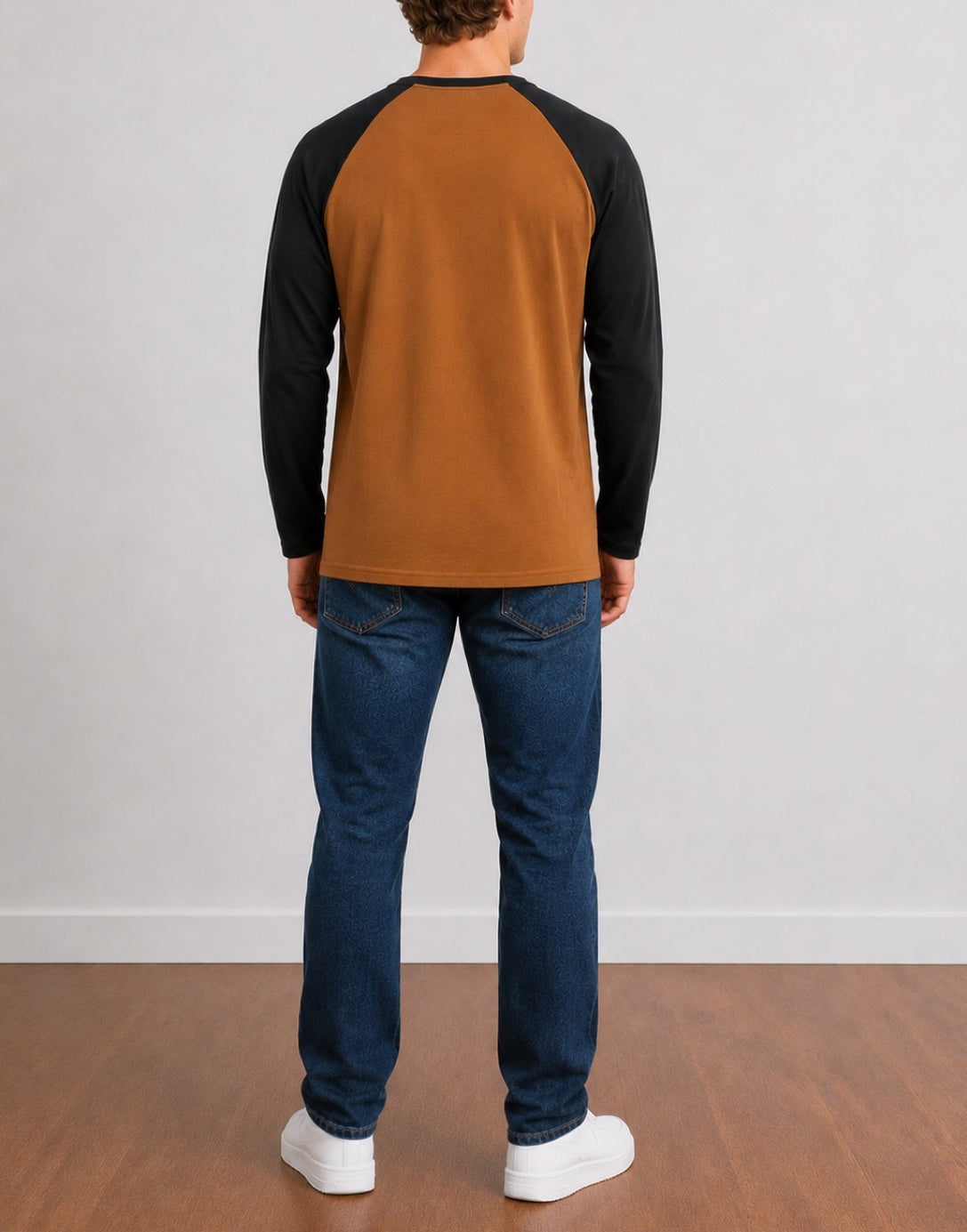 Back view of a man wearing a brown and black Raglan Long Sleeve Henley Top, dark blue jeans, and white sneakers, standing against a light gray wall and wood floor.