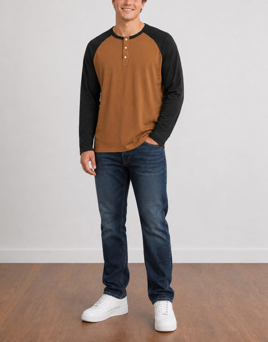 A man wearing a brown and black Raglan Long Sleeve Henley Top, dark blue jeans, and white sneakers, standing against a light gray wall and wood floor.