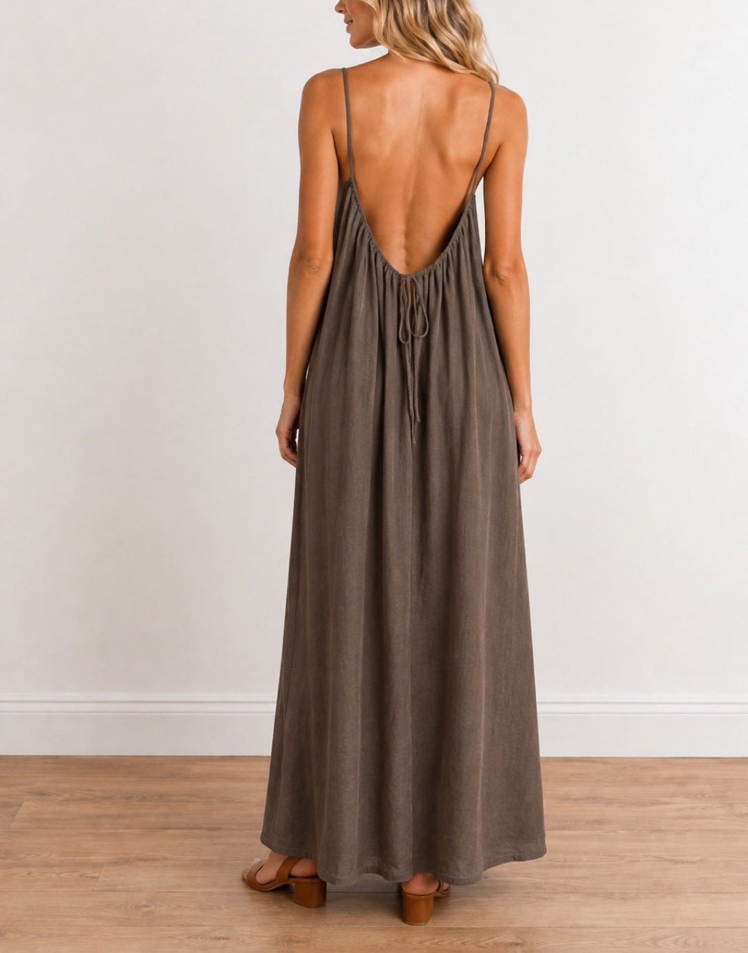 A woman viewed from the back wearing a long, dark brown Maxi Dress with thin straps and a deep open back, standing on a light wood floor against a white wall.