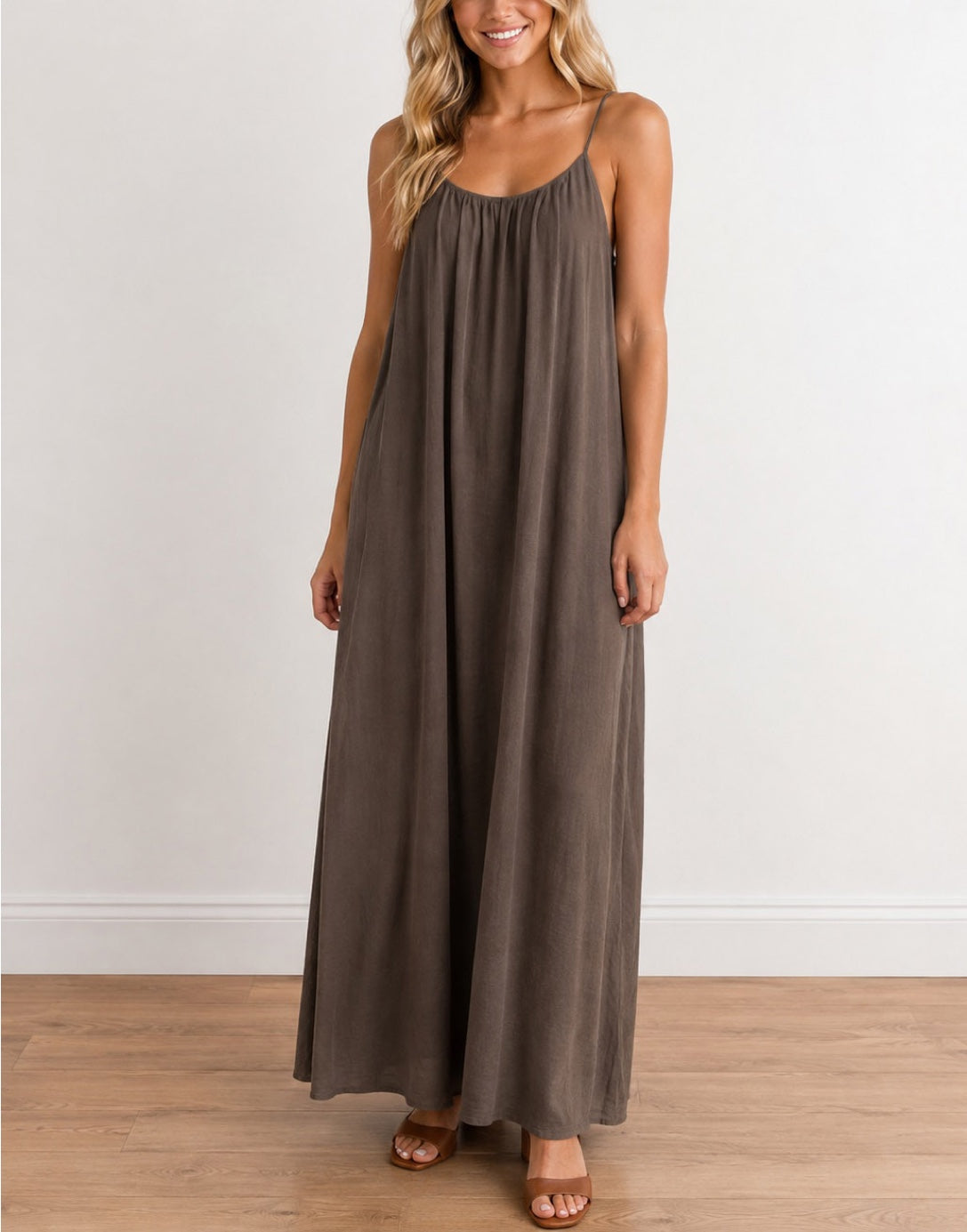 A woman wearing a Sleeveless, Floor-Length Maxi Dress in a muted taupe-brown color, standing against a plain white wall on a light wood floor.
