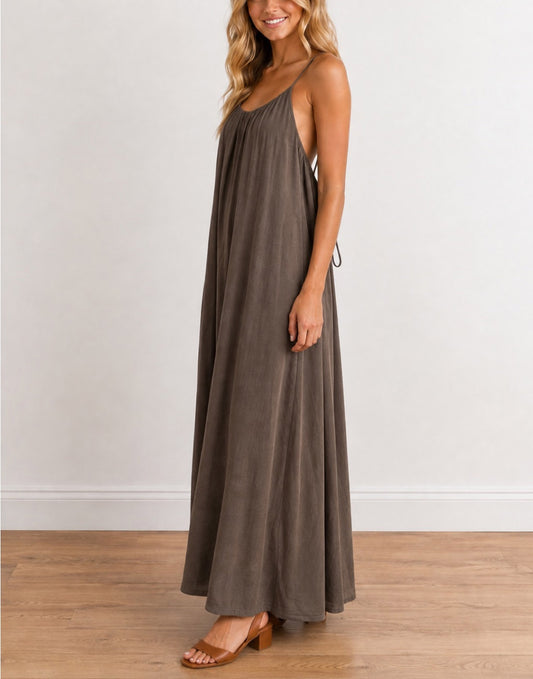 Side view of a woman modeling a Long, Sleeveless Maxi Dress in a taupe brown color, standing on a light wood floor against a white wall.