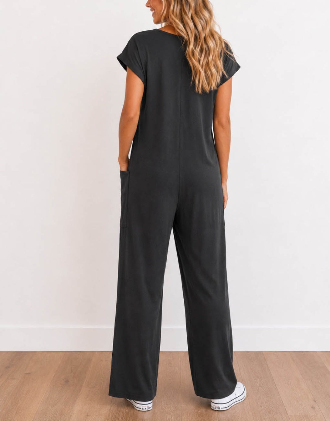 Back view of woman wearing a charcoal gray Oversized Knit Tee Jumpsuit with white shoes on a wooden floor with a plain background.