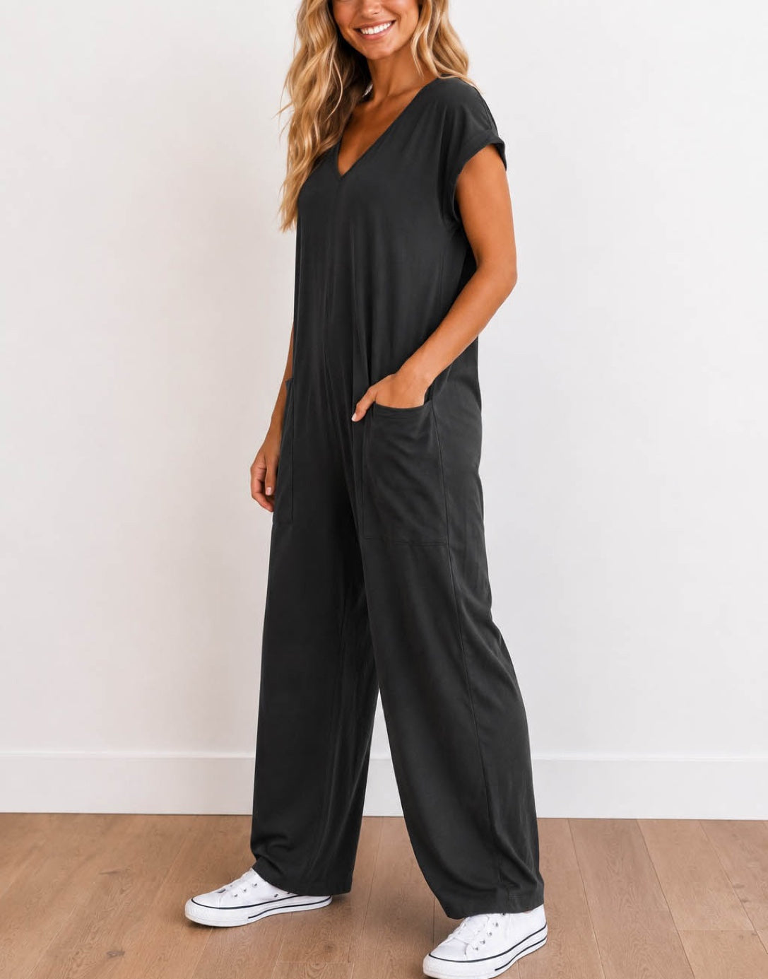 Side view of woman wearing a charcoal gray Oversized Knit Tee Jumpsuit with white shoes on a wooden floor with a plain background.
