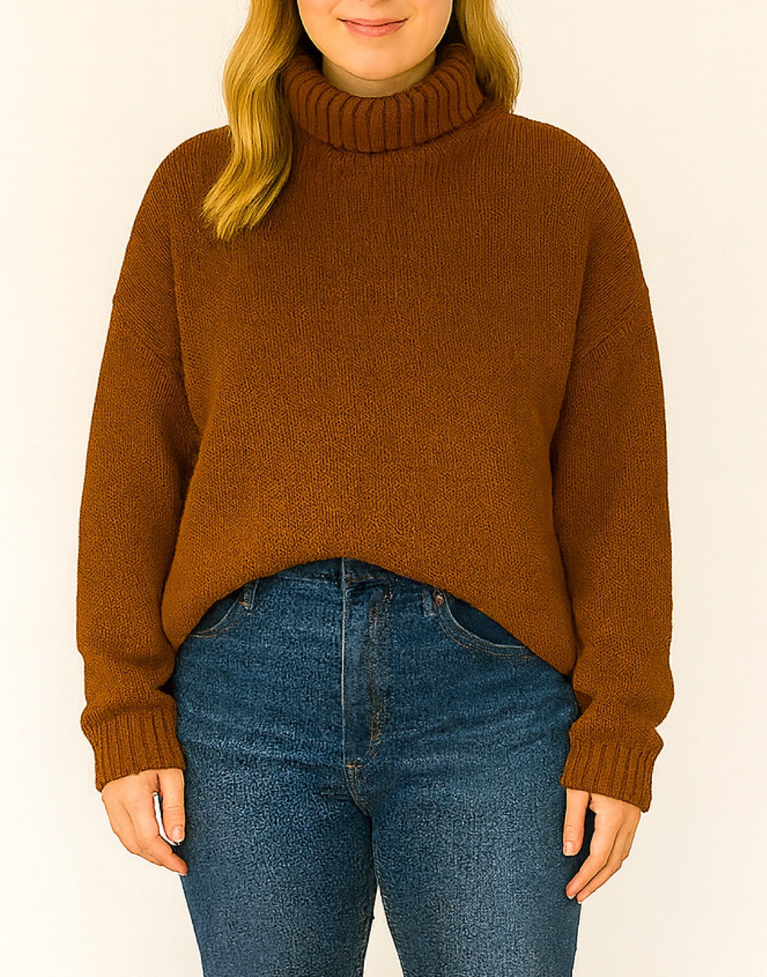 Person wearing a brown oversized turtleneck sweater and blue jeans on a plain background