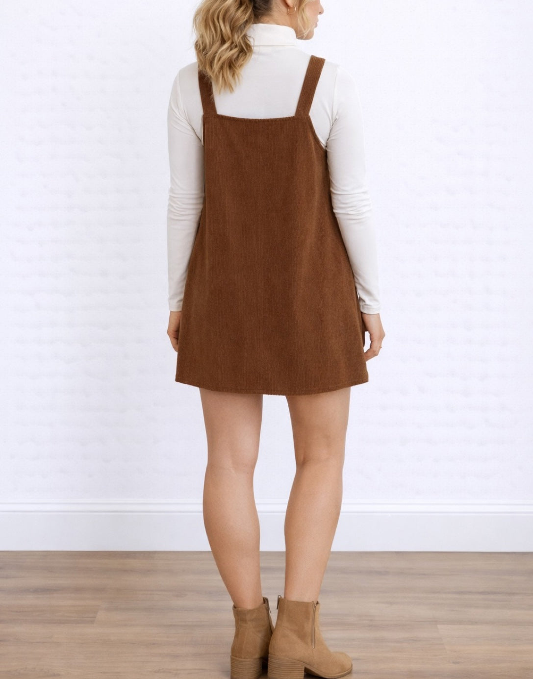 Back view of woman wearing a brown Pinafore Mini Dress over a white long-sleeve shirt on a white background.