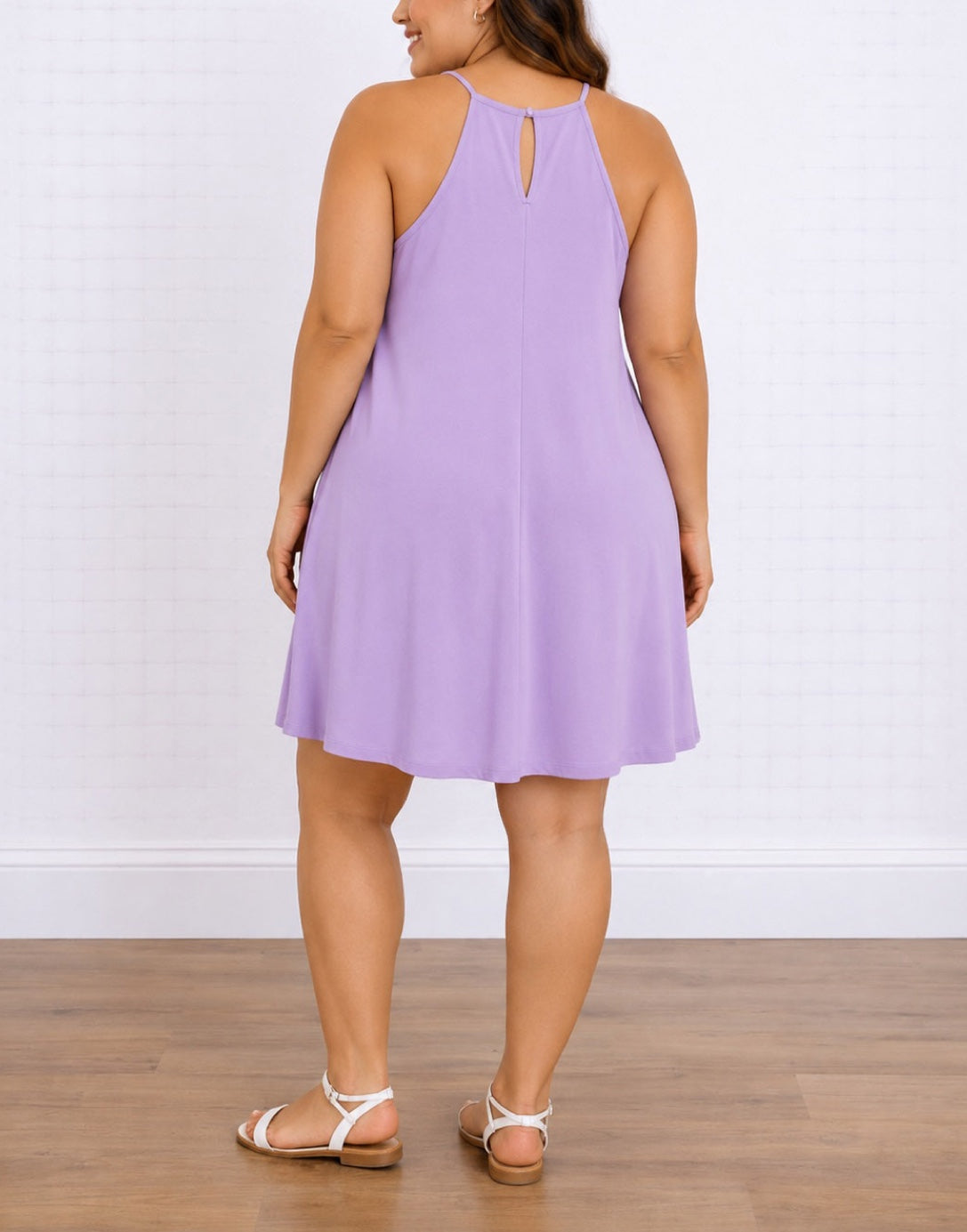 A woman shown from the back wearing a sleeveless lavender-purple Plus Size Halter Fit-and-Flare dress with a small keyhole cutout at the back, paired with white strappy flat sandals, standing on a wooden floor against a white tiled wall.