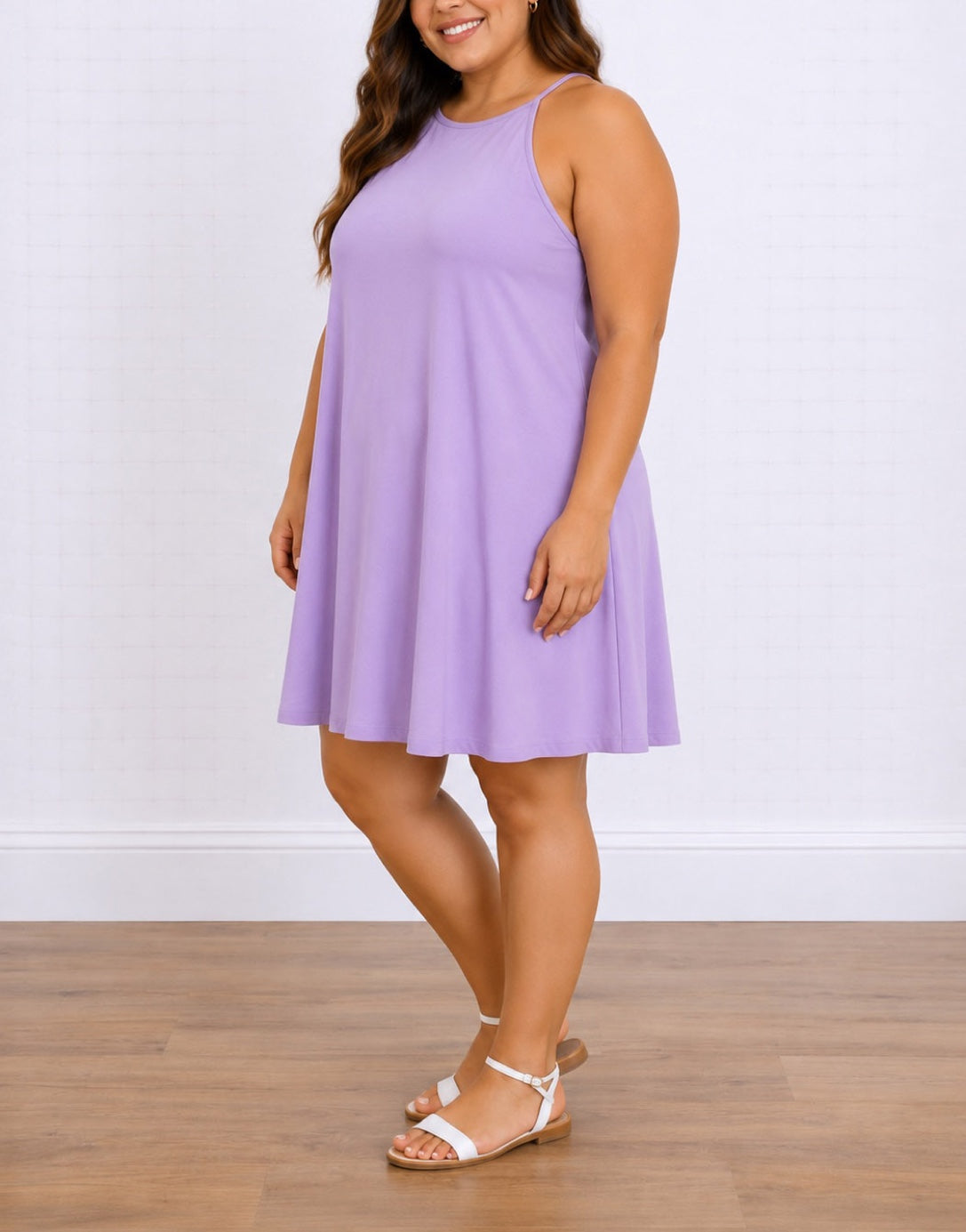 Side view of a woman modeling a sleeveless lavender-purple Plus Size Halter A-line mini dress with a high neckline, paired with white strappy flat sandals, standing against a white brick wall and wood floor.