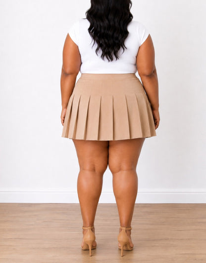 Back view of a woman modeling a beige Plus Size High Waist Pleated Mini Skirt paired with a white sleeveless top and tan heeled sandals, standing against a white wall and light wood floor.