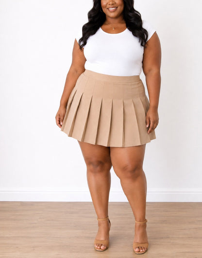 A woman modeling a beige Plus Size High Waist Pleated Mini Skirt paired with a white sleeveless top and tan heeled sandals, standing against a white wall and light wood floor.