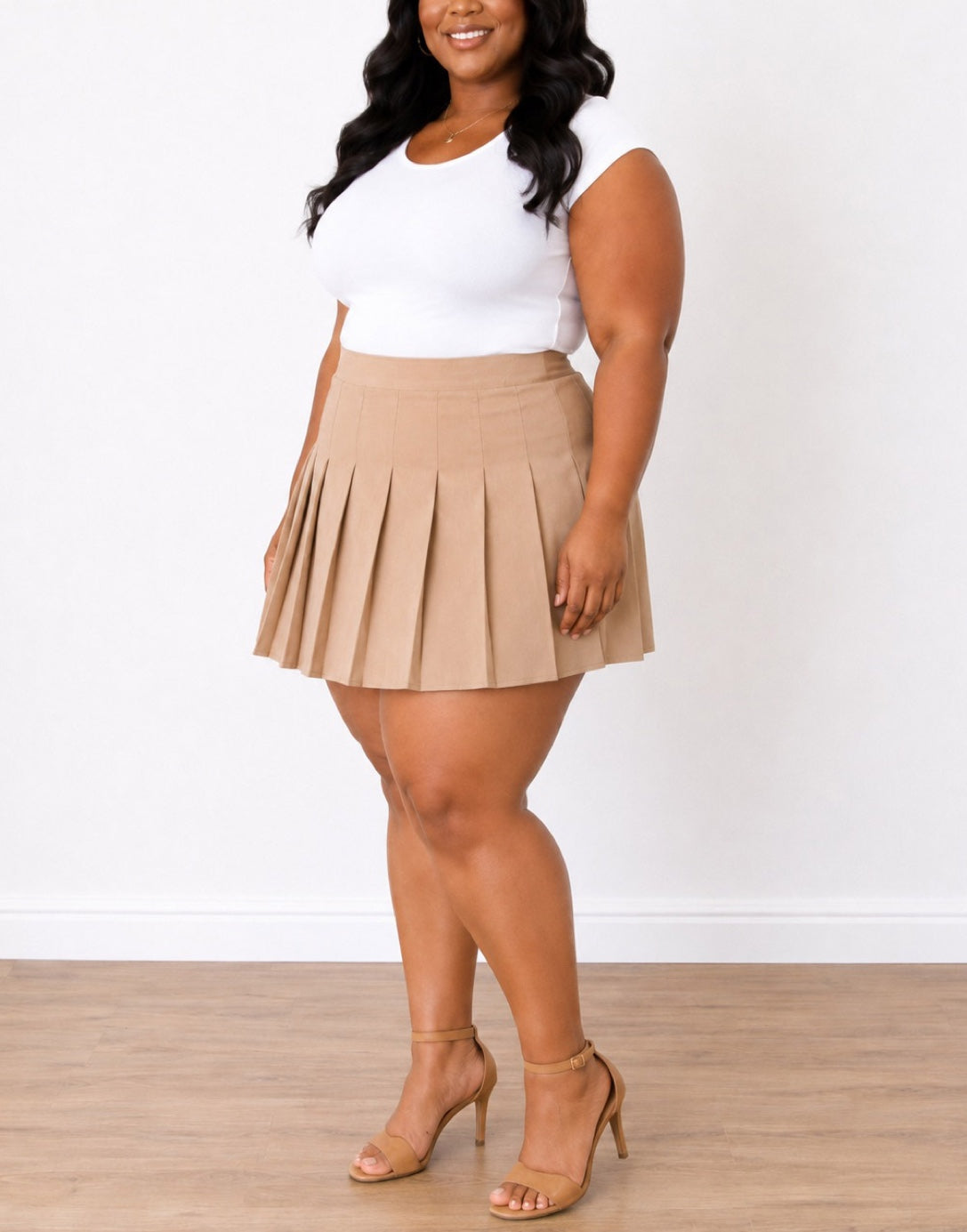 Side view of a woman modeling a beige Plus Size High Waist Pleated Mini Skirt paired with a white sleeveless top and tan heeled sandals, standing against a white wall and light wood floor.