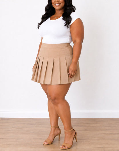 Side view of a woman modeling a beige Plus Size High Waist Pleated Mini Skirt paired with a white sleeveless top and tan heeled sandals, standing against a white wall and light wood floor.