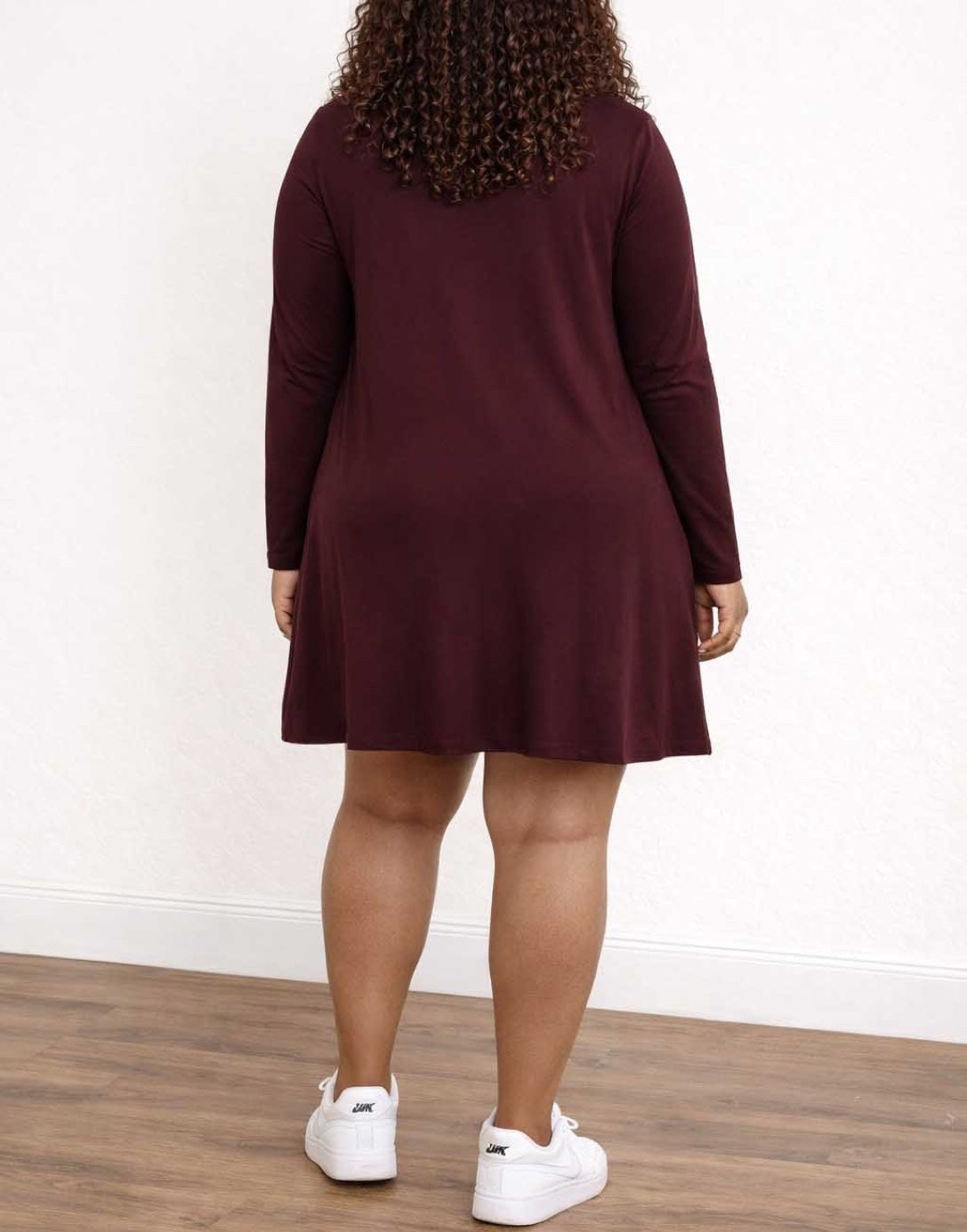 Back view of woman wearing a burgundy long-sleeve dress with white sneakers on a plain background.