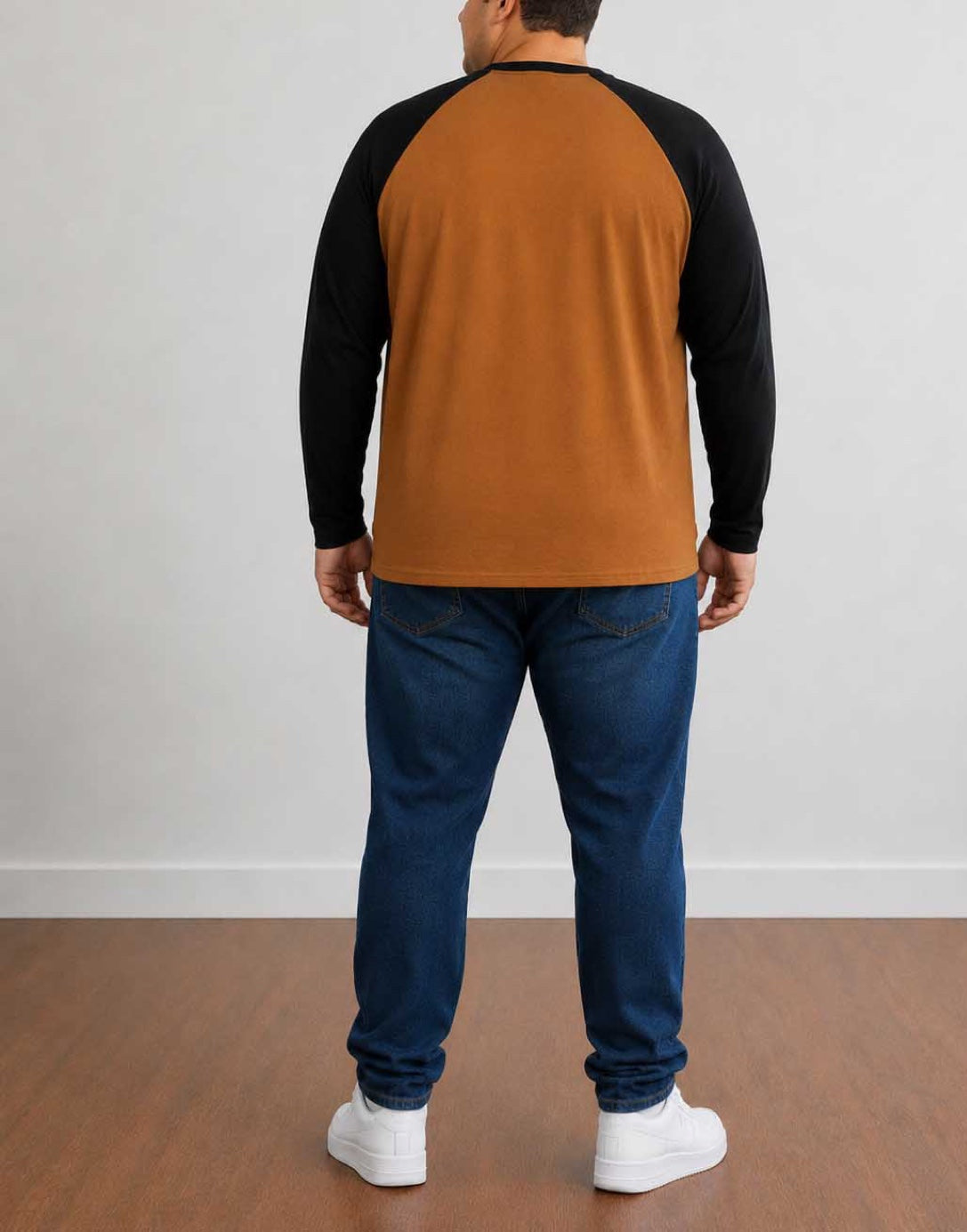 A man viewed from the back wearing a two-tone Plus Size Raglan Long-Sleeve Shirt with a mustard brown body and black sleeves, paired with blue jeans and white sneakers, standing against a plain light gray wall.