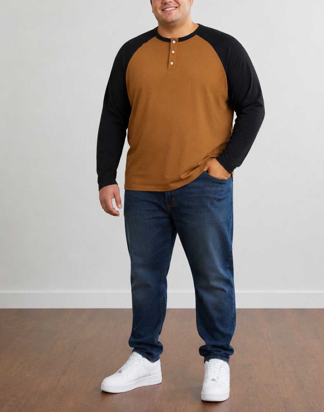 A man wearing a two-tone Plus Size Raglan Long Sleeve Henley Top with a brown body and black sleeves, paired with dark blue jeans and white sneakers, standing against a plain light gray wall.