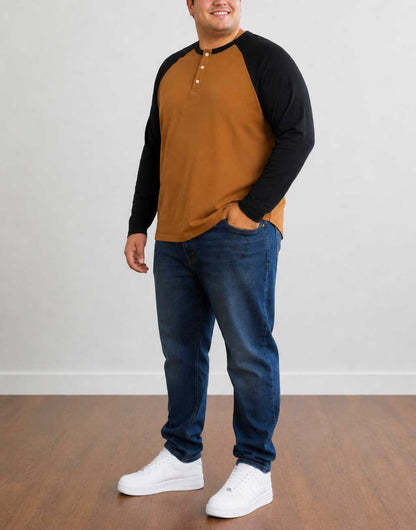 Side view of a man wearing a brown and black Plus Size Raglan Long Sleeve Henley Top, blue jeans, and white sneakers, standing against a plain light gray wall and wooden floor.