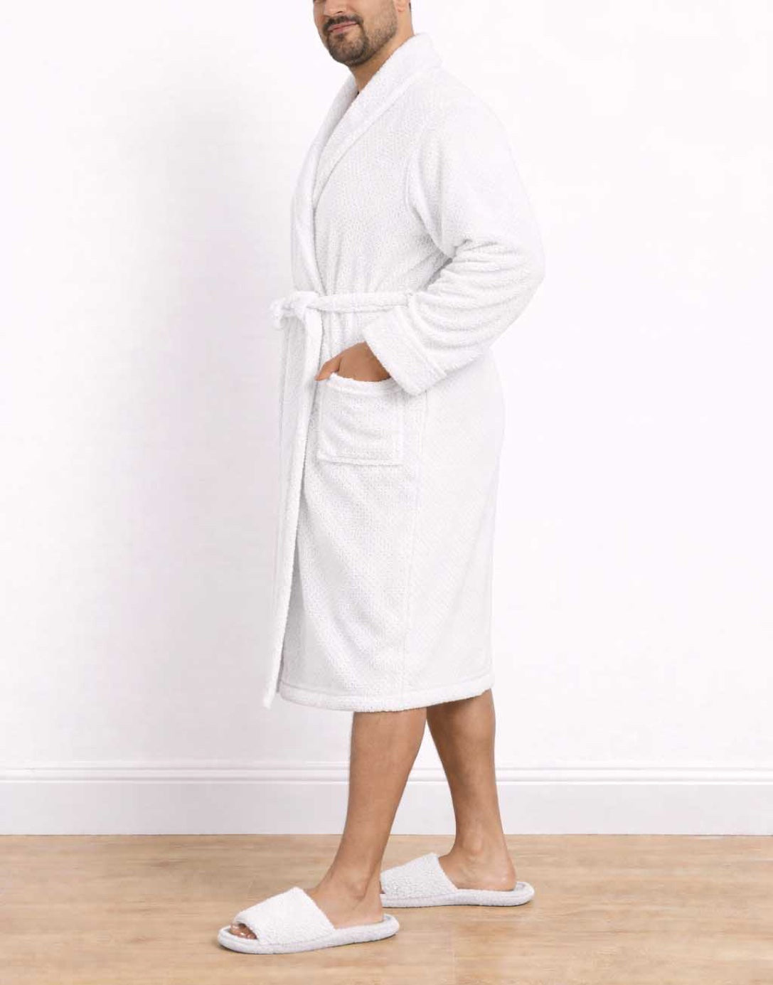 Side view of man wearing a white Plus Size Mens Shawl Collar Bathrobe and slippers on a wooden floor with a white wall background.