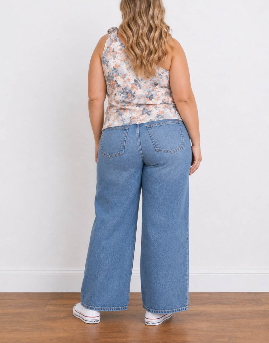 Back view of a woman modeling a floral Plus Size One Shoulder Top and wide-leg blue jeans with white sneakers, standing against a plain white wall on a wooden floor.