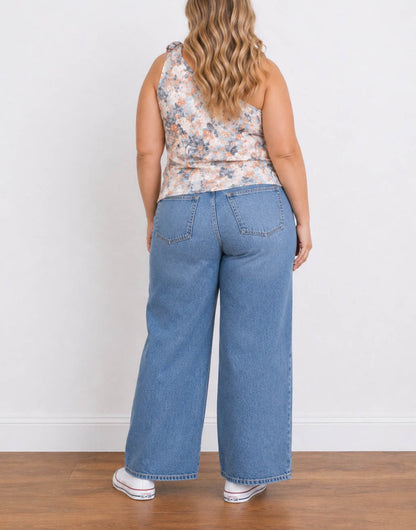 Back view of a woman modeling a floral Plus Size One Shoulder Top and wide-leg blue jeans with white sneakers, standing against a plain white wall on a wooden floor.
