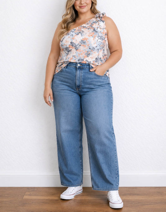 A woman wearing a sleeveless one-shoulder floral top and wide-leg blue jeans with white sneakers, standing against a white wall on a wood floor.