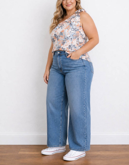 Side view of a woman modeling a floral Plus Size One Shoulder Top and wide-leg blue jeans with white sneakers, standing against a plain white wall on a wooden floor.