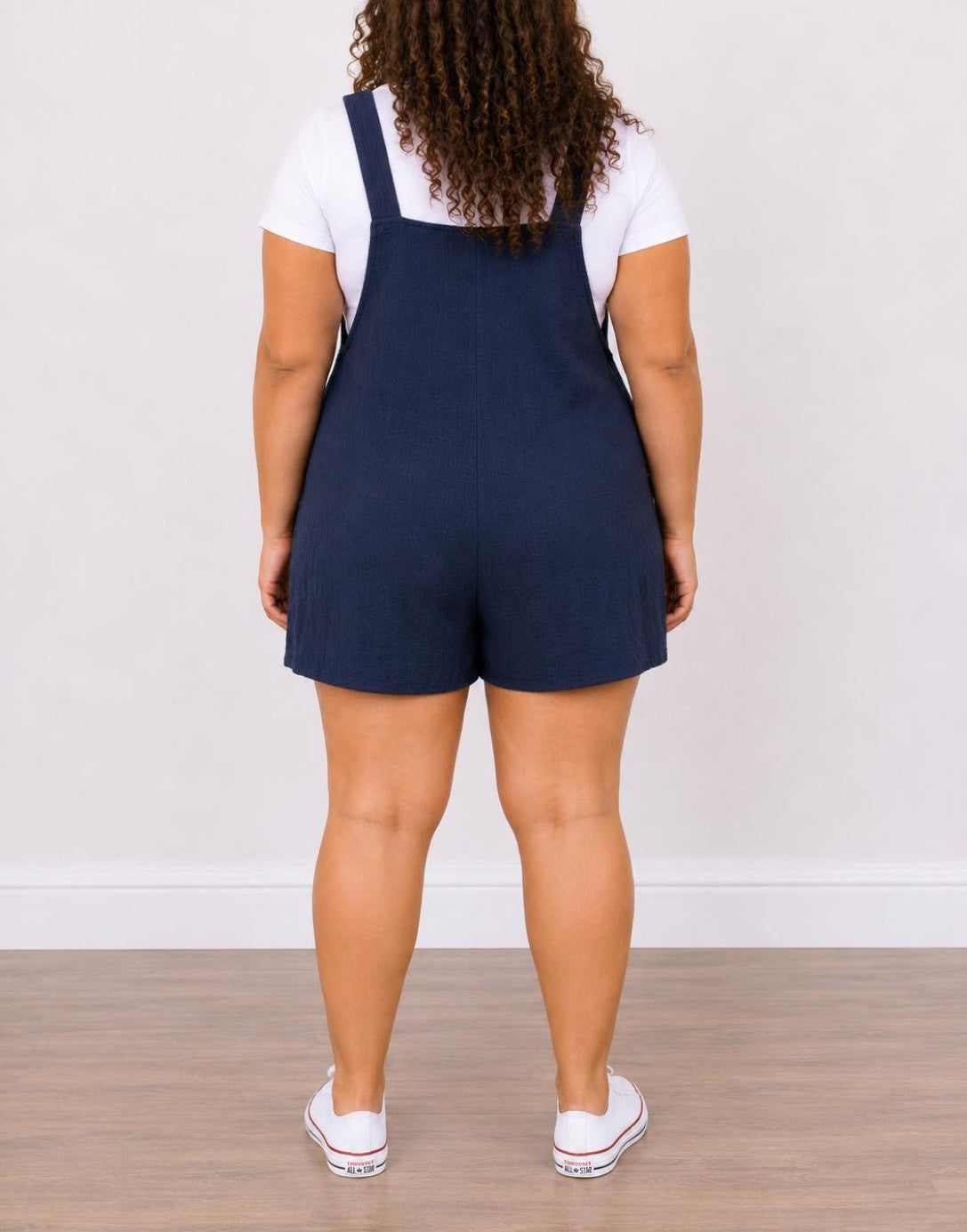 Back view of woman wearing navy blue Plus Size Overall Romper with a white shirt underneath on a plain background.