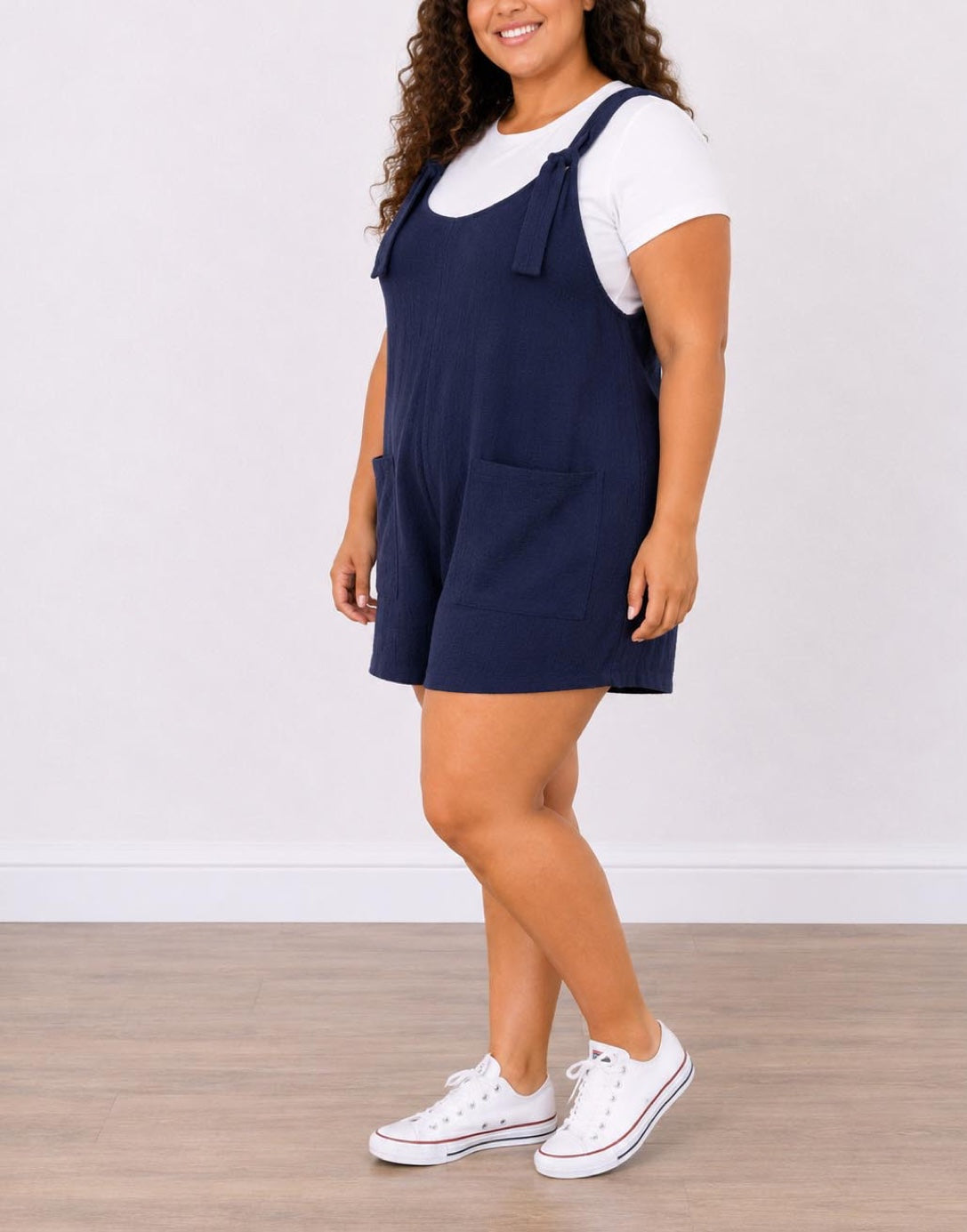 Side view of woman wearing navy blue Plus Size Overall Romper with a white shirt underneath on a plain background.