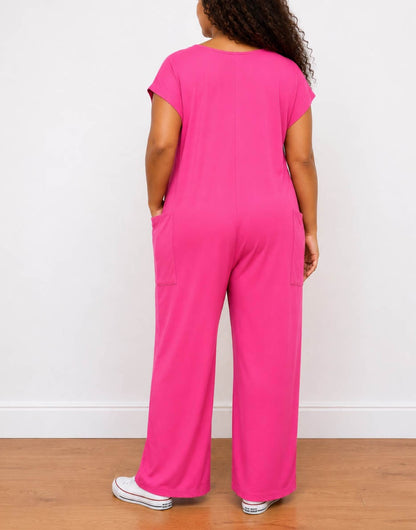 A woman viewed from behind wearing a bright pink Plus Size Short-Sleeve Oversized Knit Jumpsuit with wide legs and white sneakers, standing against a white wall on a wooden floor.