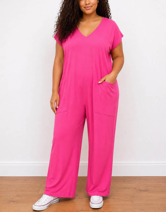 A woman modeling a bright pink Plus Size Oversized Knit Tee Jumpsuit with a V-neck and wide legs, standing on a wooden floor against a white wall.