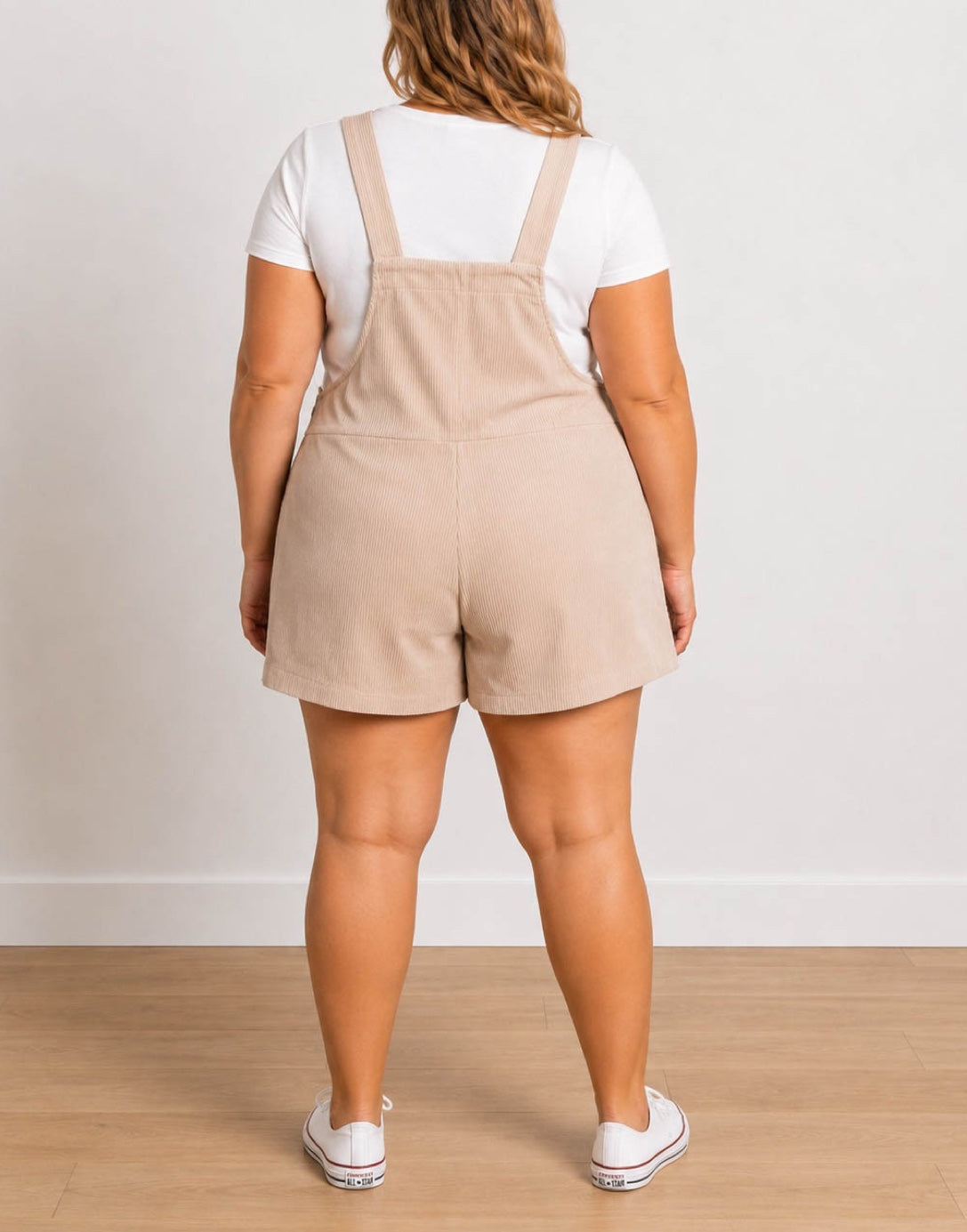 Back view of a woman wearing a beige corduroy Plus Size Overall Romper over a white t-shirt, paired with white sneakers, standing against a neutral light gray wall and wooden floor.