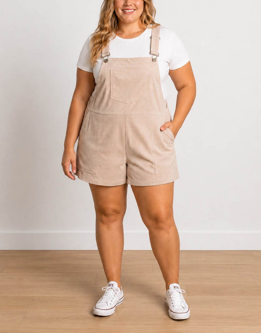 A woman wearing a beige corduroy Plus Size Pocket Overall Romper over a white t-shirt, paired with white sneakers, standing against a neutral light gray wall and wooden floor.