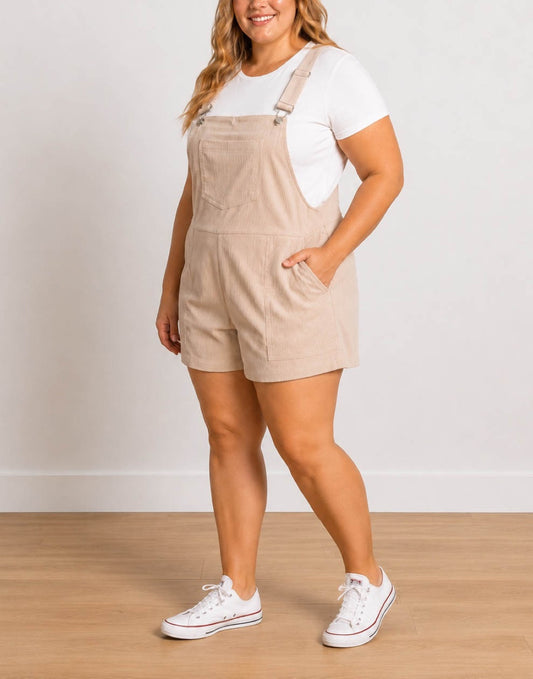 Side view of a woman wearing a beige corduroy Plus Size Pocket Overall Romper over a white t-shirt, paired with white sneakers, standing against a neutral light gray wall and wooden floor.