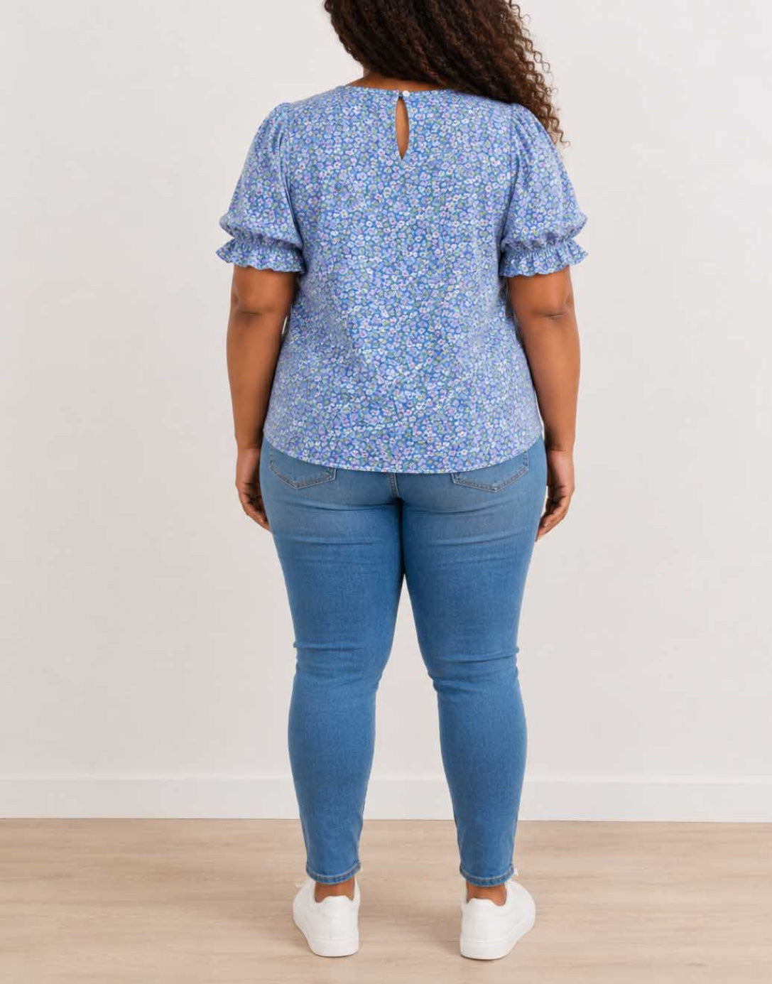 A woman viewed from the back wearing a light blue and white floral print Plus Size Short-Sleeve Top with Ruffled Sleeves, blue skinny jeans, and white sneakers, standing against a plain off-white wall.