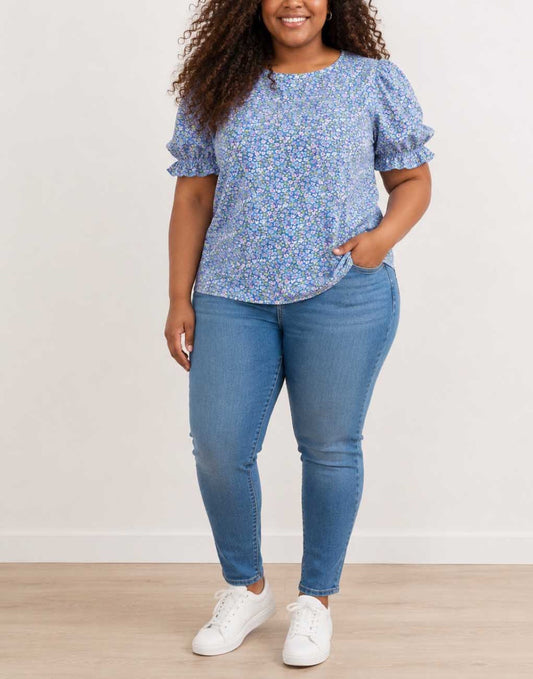 A woman modeling a light blue Plus Size Puff Sleeve Blouse with a dense small floral print, paired with medium-wash blue jeans and white sneakers, standing against a plain off-white wall.