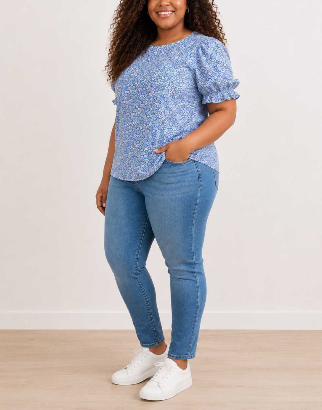 Side view of a woman modeling a light blue floral Plus Size Short-Sleeve Top with Puff Sleeves, paired with blue jeans and white sneakers, standing against a plain off-white wall.