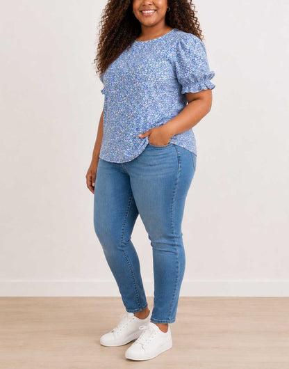 Side view of a woman modeling a light blue floral Plus Size Short-Sleeve Top with Puff Sleeves, paired with blue jeans and white sneakers, standing against a plain off-white wall.