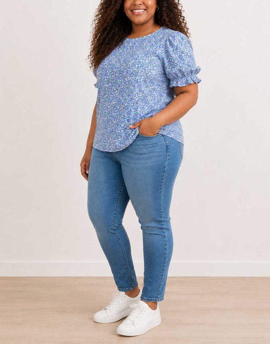 Side view of a woman modeling a light blue floral Plus Size Short-Sleeve Top with Puff Sleeves, paired with blue jeans and white sneakers, standing against a plain off-white wall.