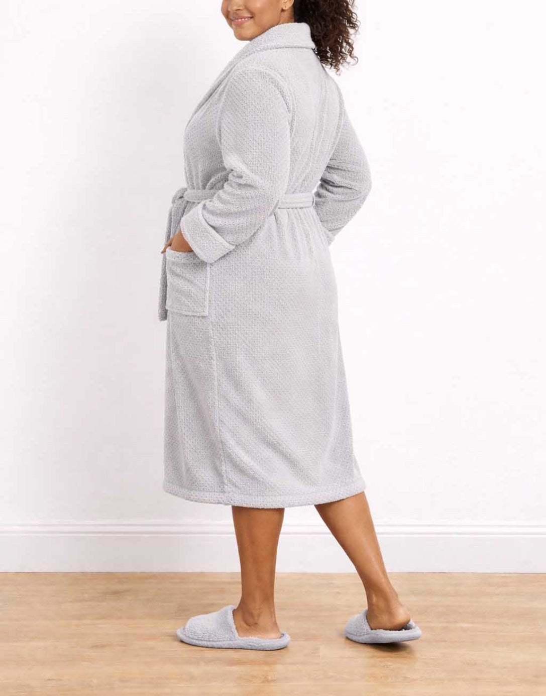 Side view of woman wearing a gray Plus Size Shawl Collar Bathrobe and slippers on a wooden floor with a white wall background.