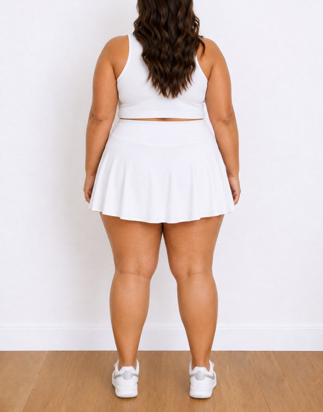 A plus-size woman shown from the back wearing a white sleeveless crop top and matching white Pleated Sport Skort, paired with white sneakers, standing against a white wall on a light wood floor.