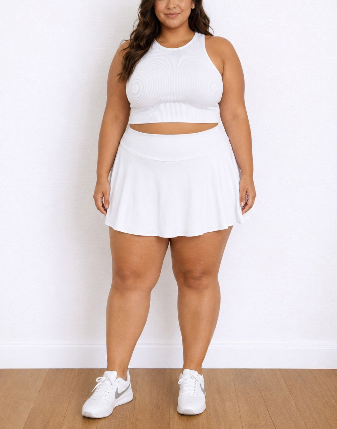 A plus-size woman modeling a white sleeveless crop top and matching white Sport Skort, paired with white sneakers, standing against a white wall on a light wood floor.