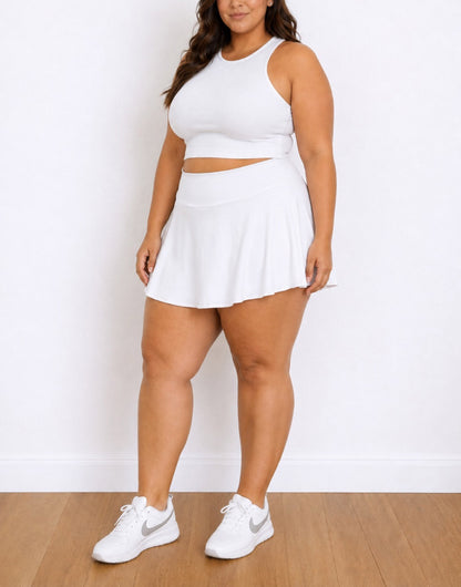 A woman modeling a white two-piece athletic set consisting of a sleeveless crop top and a Plus Size High-Waisted Sport Skort, paired with white sneakers, standing against a white wall and wood floor.