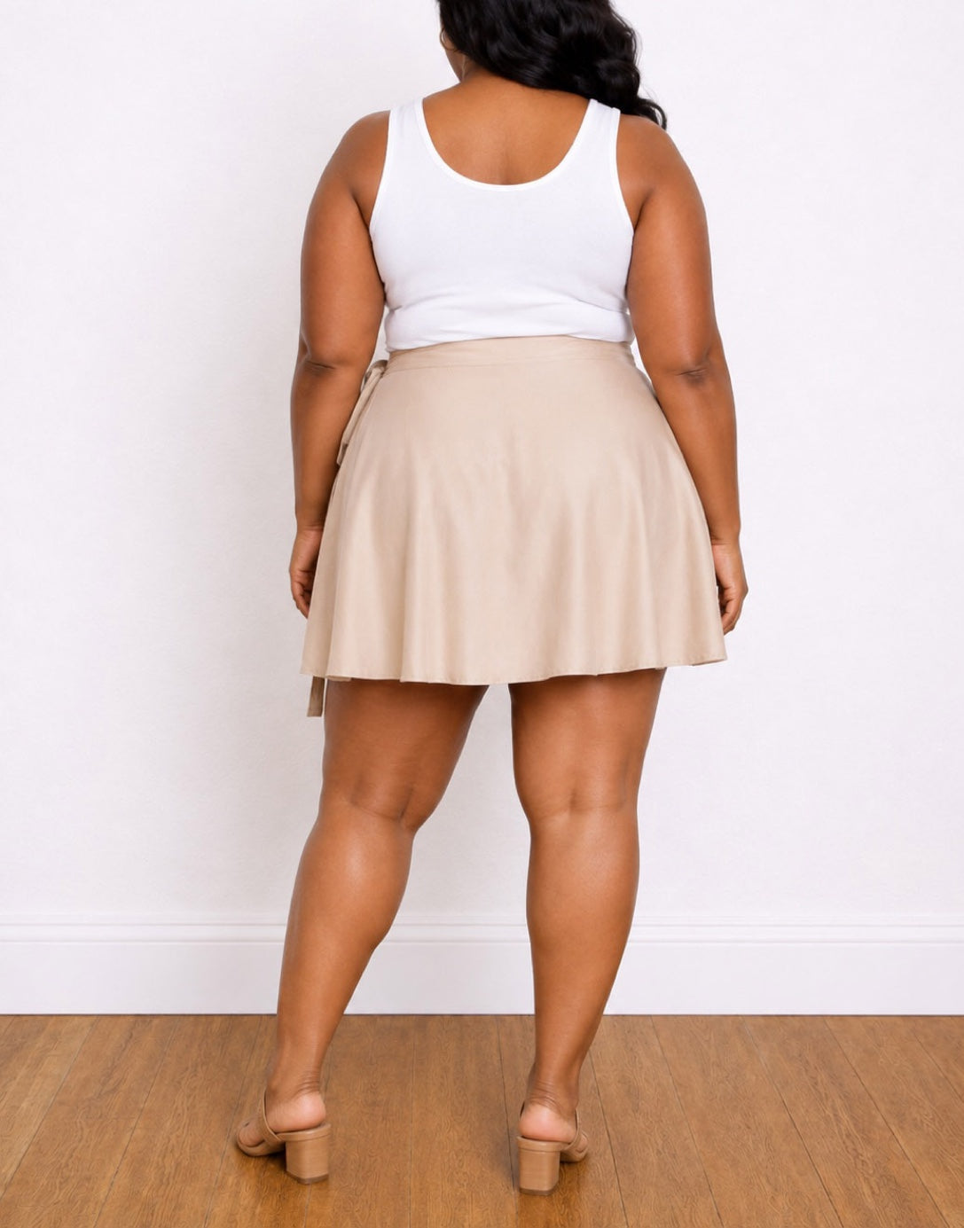 A woman shown from the back wearing a white tank top and a beige Plus Size Flared Mini Skirt, standing on a wooden floor against a white wall.