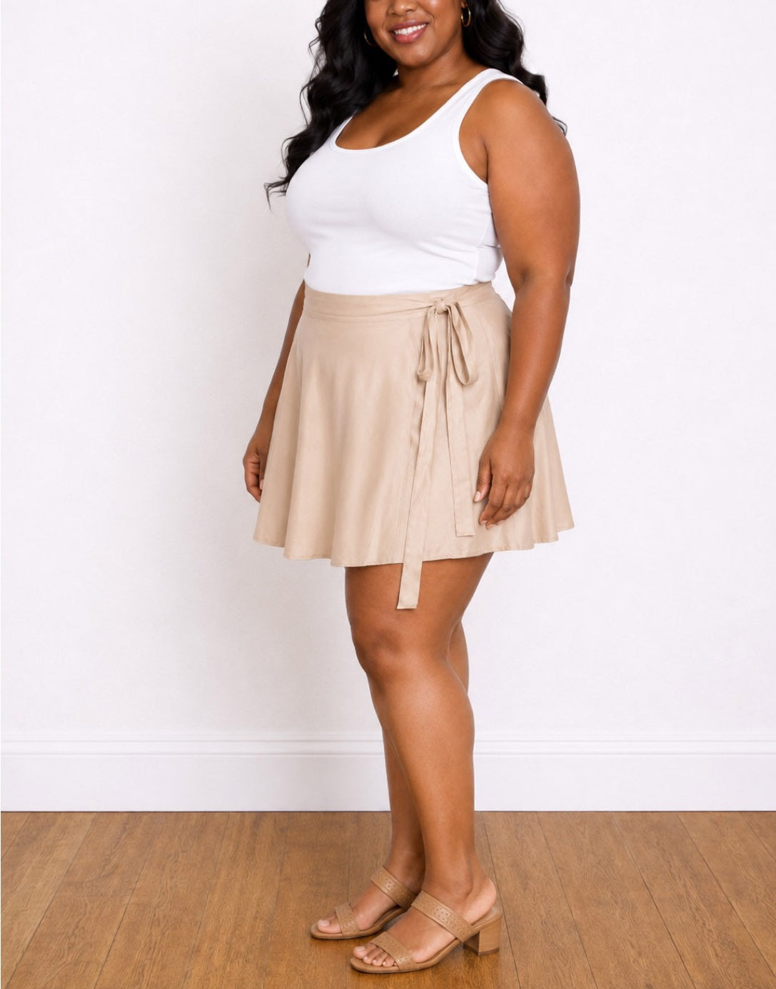 A woman modeling a beige Plus Size Wrap-Style Mini Skirt with a tie waist, paired with a white tank top and beige heeled sandals, standing against a white wall and wood floor.
