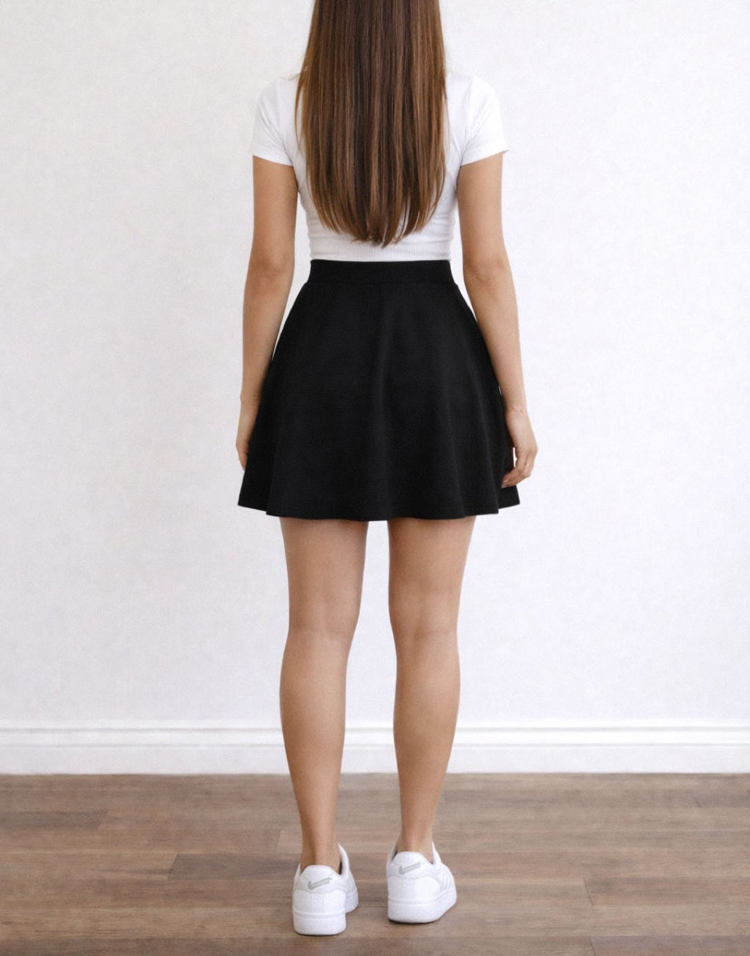 Back view of woman wearing a black Skater Knit Skirt and white top in a plain room with wooden floor.